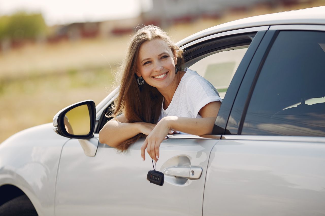 Happy female driver in white t shirt leaning out of side door window with car key in hand and looking away with smile in sunny summer day